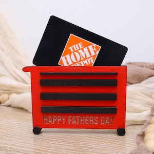 May include: A red wooden toolbox-shaped gift card holder with black accents and the text "HAPPY FATHERS DAY." A black gift card with the Home Depot logo is inserted into the holder. The holder has small black wheels.