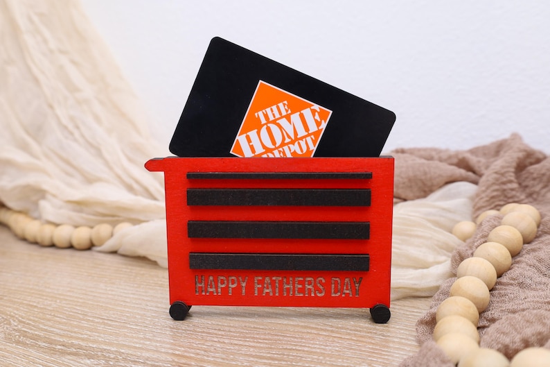May include: A red wooden toolbox-shaped gift card holder with black accents and the text "HAPPY FATHERS DAY." A black gift card with the Home Depot logo is inserted into the holder. The holder has small black wheels.