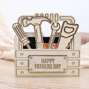 May include: A wooden toolbox-shaped gift box with cut-out tools, including a screwdriver, hammer, wrench, and saw. The box has the words "HAPPY FATHERS DAY" engraved on the front. The box is made of light-colored wood.