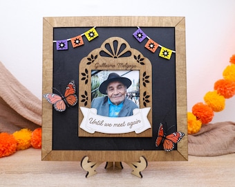 May include: Wooden memorial frame with a photo of a man wearing a hat, adorned with butterfly accents and a banner that reads "Until we meet again." The frame includes decorative paper flags and a wooden stand.