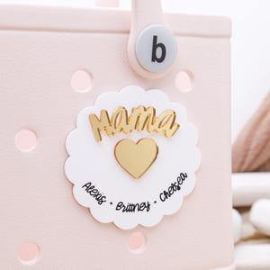 May include: A pink tote bag with a white circle sticker that says "Mama" in gold letters with a gold heart in the center. The sticker also says "Alexis - Brittney - Chelsea" around the heart.