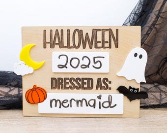 May include: Wooden Halloween sign with the words "HALLOWEEN 2025 DRESSED AS: mermaid". The sign has decorative cutouts of a yellow moon, white cloud, orange pumpkin, white ghost, and black bat.