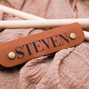 May include: A brown leather strap with the name "STEVEN" embossed in black lettering. The strap has two brass rivets.