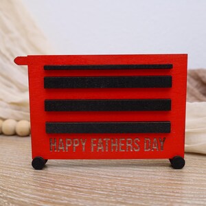 May include: A red wooden toolbox with black accents and the words "HAPPY FATHERS DAY" cut into the front. The toolbox has four black wheels and three black horizontal bars. The background includes a beige fabric and wooden beads.
