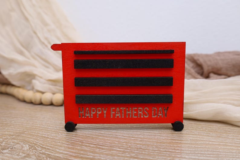 May include: A red wooden toolbox with black accents and the words "HAPPY FATHERS DAY" cut into the front. The toolbox has four black wheels and three black horizontal bars. The background includes a beige fabric and wooden beads.