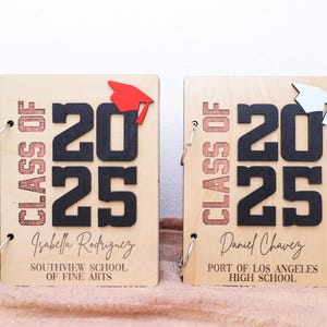 May include: Two wooden graduation photo albums with black numbers 2025 and the text "Class of" in a brown color. The albums have a red and white graduation cap on the front. The text "Isabella Rodriguez" and "Southview School of Fine Arts" is on the left album. The text "Daniel Chavez" and "Port of Los Angeles High School" is on the right album.