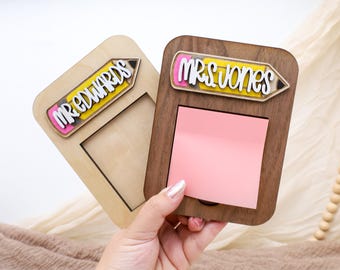May include: Two wooden memo holders, one light-colored and one dark-colored, each with a pencil-shaped nameplate. The light-colored holder has "MR EDWARDS" on it, and the dark-colored holder has "MRS JONES" on it. A pink sticky note pad is in the dark-colored holder.
