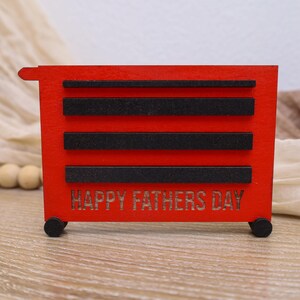 May include: A red wooden toolbox-shaped decoration with black accents and the words "HAPPY FATHERS DAY" cut out on the front. The decoration has small black wheels and is designed to resemble a miniature tool chest.