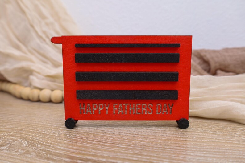 May include: A red wooden toolbox-shaped decoration with black accents and the words "HAPPY FATHERS DAY" cut out on the front. The decoration has small black wheels and is designed to resemble a miniature tool chest.
