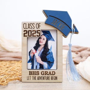 May include: Wooden photo frame with a graduation theme. The frame holds a photo of a graduate in a blue cap and gown. A blue mortarboard with the name "Jackie" is attached to the frame, along with a light blue tassel. The frame reads "Class of 2025" and "BHS Grad Let the Adventure Begin."