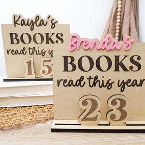 May include: Wooden book tracker signs with the names "Kayla" and "Brenda" in pink. The signs display the text "BOOKS read this year" and the numbers 15 and 23. The signs are made of light-colored wood.