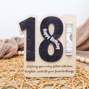 May include: Wooden birthday card with the number 18 in black glitter. The number 8 has "Happy Birthday" in white text. A slot holds a rolled-up bill. The bottom of the card has a message: "Wishing you a day filled with love, laughter, and all your favorite things."