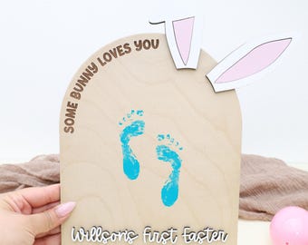 Op de afbeelding: Houten paasbord met konijnenoren en de tekst "Some Bunny Loves You". Het heeft blauwe voetafdrukken en de woorden "Willson's first Easter 2026". Een hand houdt het bord vast, met een roze paasei op de achtergrond.