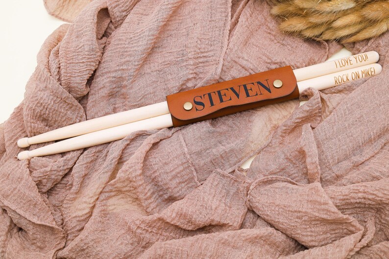 May include: A brown leather drumstick holder with the name "STEVEN" engraved on it. The holder is holding two wooden drumsticks with the text "I LOVE YOU" and "ROCK ON MIKE!" written on them.