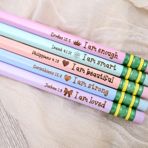 May include: Pastel-colored pencils with inspirational phrases and Bible verses. Each pencil is a different color, including pink, blue, and peach. The pencils have green and yellow striped erasers and text that reads "I am enough", "I am smart", "I am beautiful", "I am strong", and "I am loved".