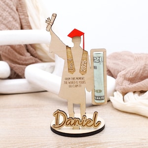 May include: Wooden graduation figure holding a diploma and money. The figure is wearing a red cap and gown and has the text "Class of 2025" on the front. The base of the figure has the text "From this moment the world is yours go claim it!" and the name "Daniel".