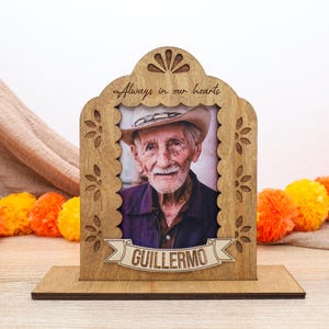 May include: A wooden memorial photo frame with a scalloped edge and laser-cut floral details. The frame features the engraved text Always in our hearts at the top and a banner at the bottom with the name GUILLERMO, holding a portrait of an older man.