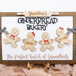 May include: A wooden sign with the text "Grandma's Gingerbread Bakery" and a gingerbread person banner with the names "Peter", "Arianna", "Jason", and "Victoria". The sign reads "The Perfect Batch of Grandkids".