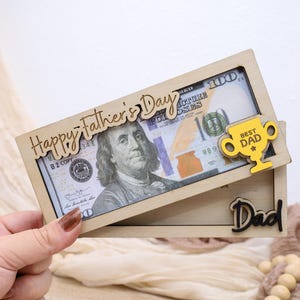 May include: A wooden Father's Day gift featuring a $100 bill framed by laser-cut text and a trophy. The top frame reads "Happy Father's Day" and includes a yellow trophy that says "Best Dad". The bottom frame says "Dad".