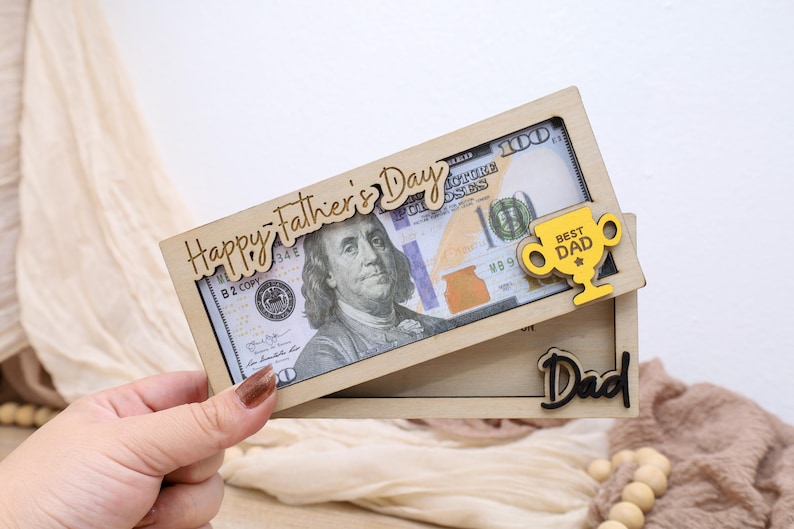 May include: A wooden Father's Day gift featuring a $100 bill framed by laser-cut text and a trophy. The top frame reads "Happy Father's Day" and includes a yellow trophy that says "Best Dad". The bottom frame says "Dad".