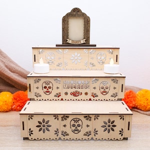 May include: A three-tiered wooden Day of the Dead altar with laser-cut skull and floral designs. The altar features a decorative frame, two battery-operated candles, and the text "Dia de los Muertos". Orange and yellow pom-poms are in the foreground.