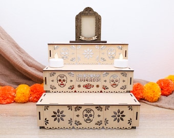 May include: A three-tiered wooden Day of the Dead altar with laser-cut skull and floral designs. The altar features a decorative frame, two battery-operated candles, and the text "Dia de los Muertos". Orange and yellow pom-poms are in the foreground.