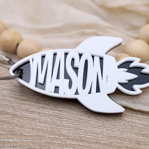 May include: A white rocket-shaped name tag with the name "MASON" cut out in white letters. The rocket has black accents and a silver-colored wire attached. The background includes wooden beads and a beige fabric.
