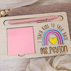May include: Wooden desk organizer with a pink pen, a pink sticky note pad, and a rainbow design with the text "I teach kids to talk back" and "Ms. Peyton". The organizer is made of light-colored wood.