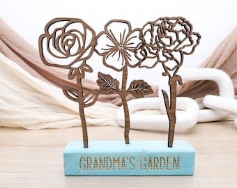 Op de afbeelding: Houten uitsnijding van drie bloemen met stelen, staand op een blauwe blok met de tekst "Grandma's Garden".