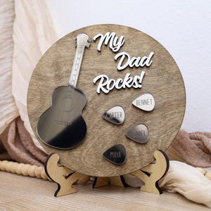 May include: A wooden guitar-themed plaque with the text "My Dad Rocks!" and guitar picks with names. The plaque features a silver guitar cutout and is supported by a wooden stand. The background includes draped fabric.