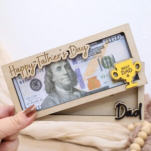 May include: A wooden Father's Day gift box with a $100 bill inside. The box has the words "Happy Father's Day" and "Best Dad" on it. The word "Dad" is also on the box.