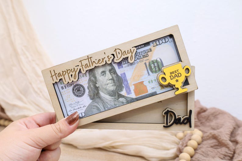 May include: A wooden Father's Day gift box with a $100 bill inside. The box has the words "Happy Father's Day" and "Best Dad" on it. The word "Dad" is also on the box.