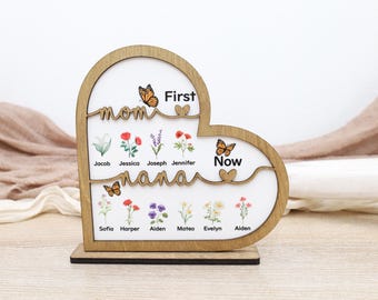 Op de afbeelding: Een houten hartvormig bord met de woorden "Mom" en "Nana" in script. Het bord bevat de woorden "First" en "Now" en is versierd met vlinders en bloemenillustraties. Namen staan onder de woorden.