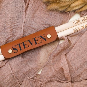 May include: A brown leather drumstick holder with the name "STEVEN" engraved on it. The holder contains two wooden drumsticks with the text "I LOVE YOU" and "ROCK ON MIKE!" engraved on them.