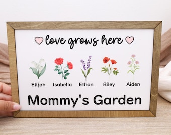 Op de afbeelding: Een ingelijst bord met een witte achtergrond en een houten frame. Het bord bevat de tekst "love grows here" met hartsymbolen en de woorden "Mommy's Garden". Het toont ook aquarel illustraties van bloemen en namen.