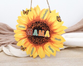 Op de afbeelding: Een houten zonnebloemvormig bord met het woord "MAMA" in kleurrijke letters. Op het bord staan de namen "James", "Isabella", "Noah", "Sophia" en "William". Er zijn ook drie oranje vlinders afgebeeld.