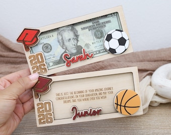 Op de afbeelding: Twee houten afstudeergeschenkdozen, een met een voetbal en de naam "Sarah", de andere met een basketbal en de naam "Junior". Elke doos heeft een afstudeerhoed, het jaar "2026" en een twintig dollar biljet.