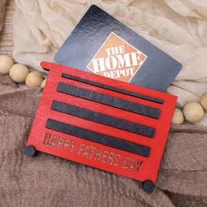 May include: A red wooden gift card holder shaped like a toolbox, with black accents and the text "HAPPY FATHERS DAY." A Home Depot gift card rests behind it. The holder has small black wheels.