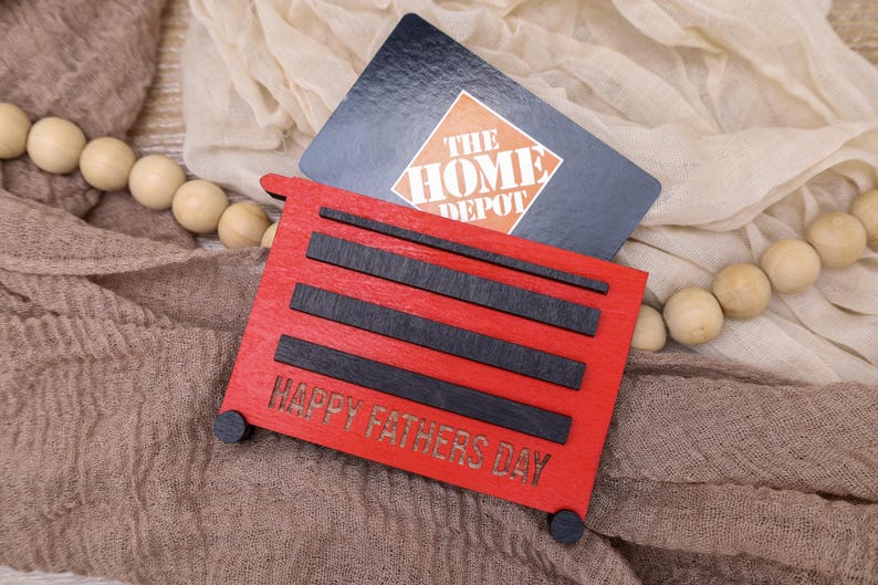 May include: A red wooden gift card holder shaped like a toolbox, with black accents and the text "HAPPY FATHERS DAY." A Home Depot gift card rests behind it. The holder has small black wheels.