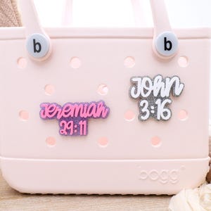 May include: A pink and white tote bag with two decorative charms. The charms are pink and purple with the text "Jeremiah 29:11" and white with the text "John 3:16".