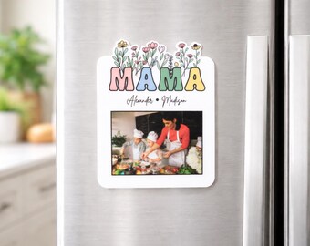 Op de afbeelding: Een witte koelkastmagneet met het woord "MAMA" in pastelkleuren, bekroond met bloemenillustraties. Hieronder wordt een familiefoto weergegeven, met de namen "Alexander & Madison" eronder geschreven. De magneet is bevestigd op een roestvrijstalen koelkast.