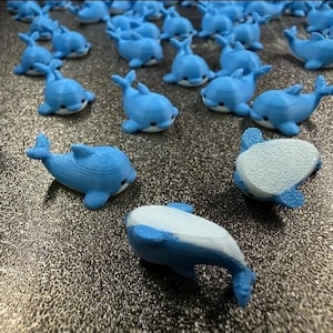 May include: A collection of small, light blue 3D-printed dolphin figurines. Each dolphin features a smooth, rounded body with a white underside, a dorsal fin, and two side fins. The figures are arranged in a group, showing both top and bottom views.