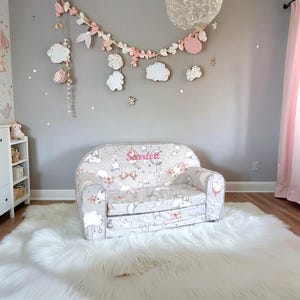 May include: A white and gray children's sofa with a floral and star pattern. The sofa has a pink embroidered name "Scarlett" on the back. The sofa is on a white fluffy rug in a room with a gray wall and a pink curtain.