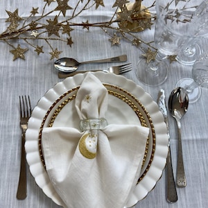 May include: Elegant table setting with white plates, gold beaded rims, and a white napkin adorned with a gold crescent moon and stars. Includes silverware, a decorative star branch, and clear glassware.