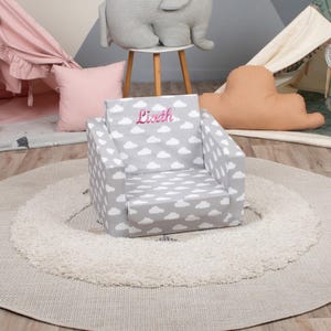 May include: A gray children's armchair with a white cloud pattern. The name "Liseth" is embroidered in pink on the backrest. The chair is on a round, light-colored rug.