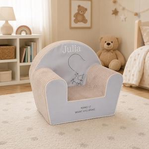 May include: A children's armchair with a light gray seat and backrest, and beige corduroy sides. The backrest features the name "Julia" and a whimsical illustration. The phrase "MAKE IT WHAT YOU MAKE" is printed on the front.