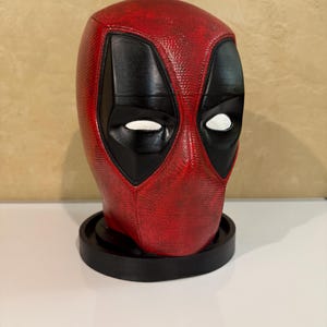May include: A red and black Deadpool head bust figurine with a textured surface. The bust is on a black circular base.