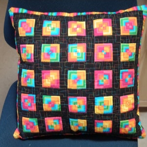 May include: A black pillow with a colorful geometric pattern. The pattern is made up of squares of different colors, including pink, orange, yellow, green, and blue. The squares are arranged in a grid pattern, and the pillow is quilted with a black thread.