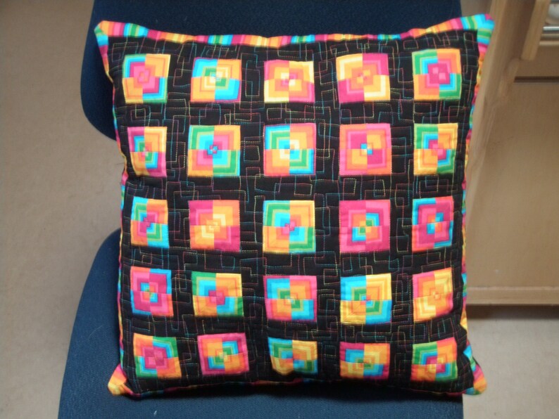May include: A black pillow with a colorful geometric pattern. The pattern is made up of squares of different colors, including pink, orange, yellow, green, and blue. The squares are arranged in a grid pattern, and the pillow is quilted with a black thread.