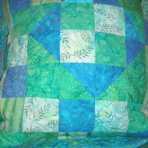 Puede incluir: Una funda de coj&iacute;n acolchada cuadrada con un dise&ntilde;o de patchwork de varios cuadrados de tela azul y verde. Los patrones de la tela incluyen colores s&oacute;lidos, motivos de hojas y texturas punteadas, unidos por costuras blancas visibles en un patr&oacute;n ondulado.